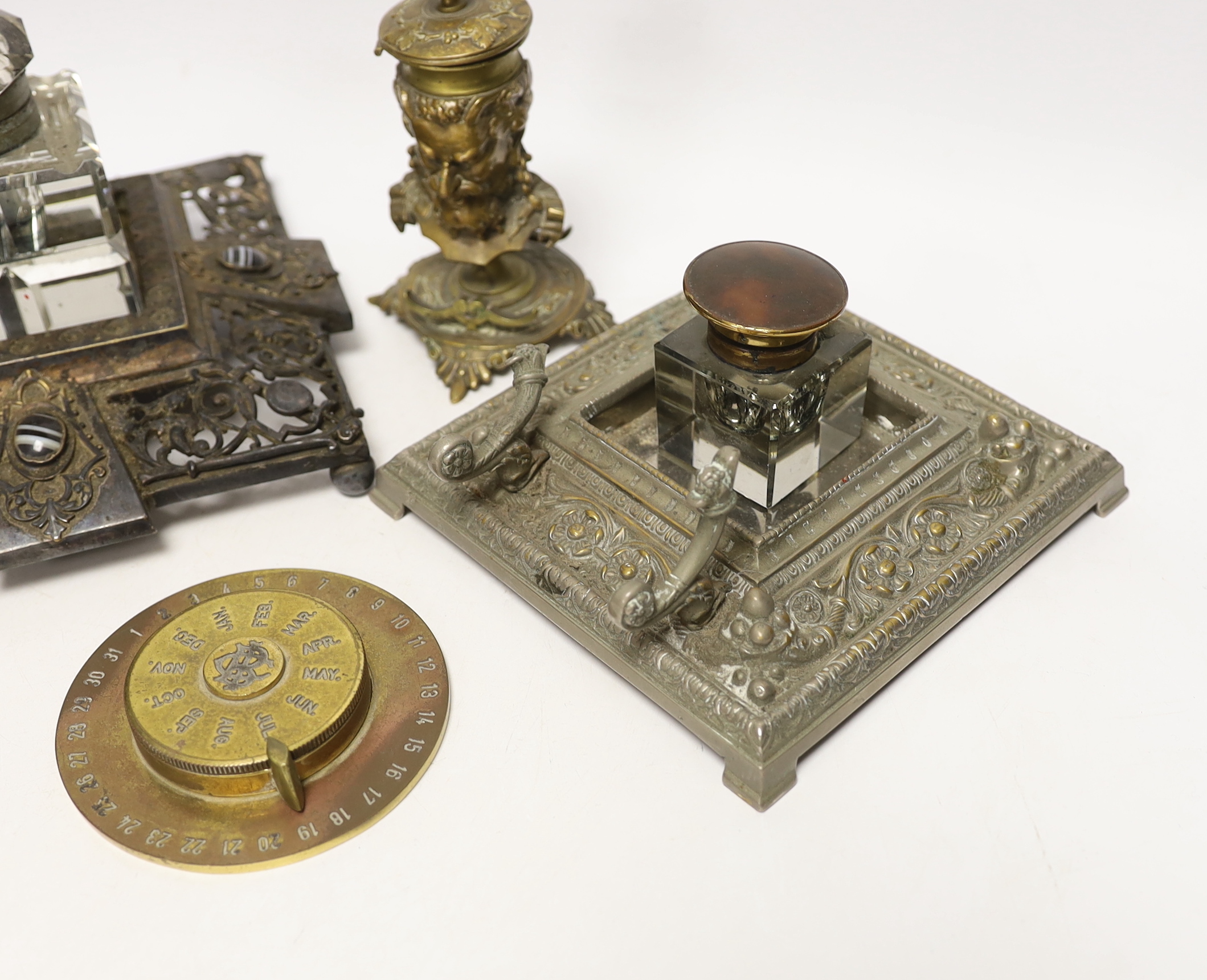 Three cast metal and cut glass ink stands, including a bronze inkwell in the form of the head of Bacus, and a brass calendar display from the Banca Commerciale Italiana, London Branch, tallest 13cm